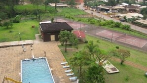 Una piscina exterior con trampolín, rodeada de césped, palmeras y una cancha de tenis.