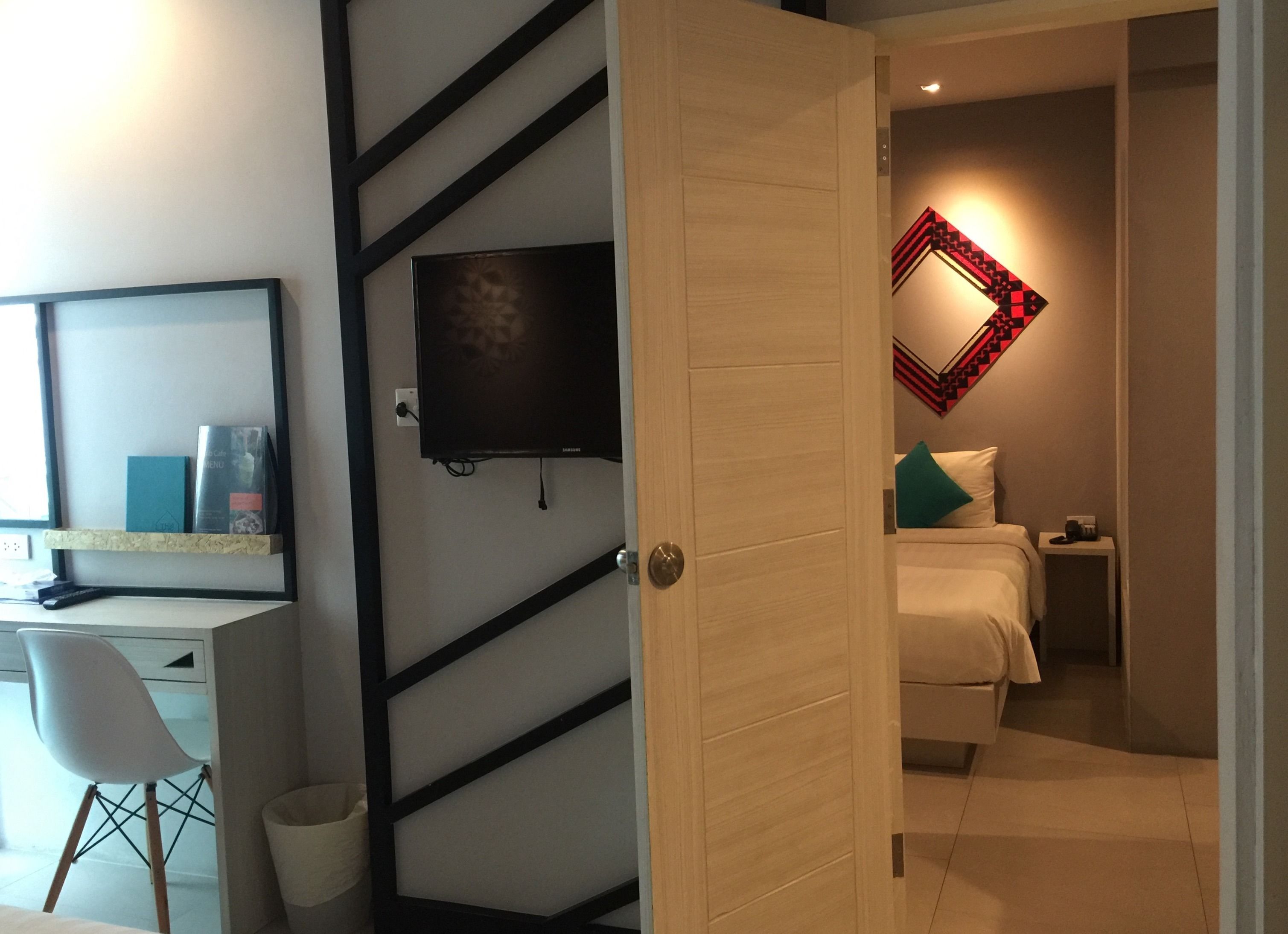 A modern hotel room with a desk, chair, TV, bed, and a decorative mirror.