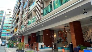 Un hôtel à plusieurs étages au design moderne, doté de balcons avec des garde-corps en verre, d’un hall d’entrée au niveau du sol avec des espaces de détente et d’une enseigne de restaurant bien visible.