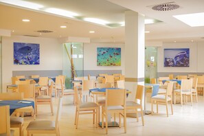 A spacious dining area with wooden tables and chairs, large windows, and artwork on the walls. - Hotel Floresta (Tías)