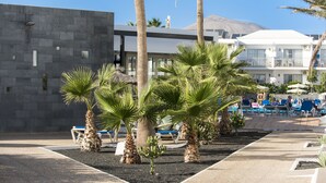 A pool area with palm trees, lounge chairs, and a modern building in the background. - Hotel Floresta (Tías)
