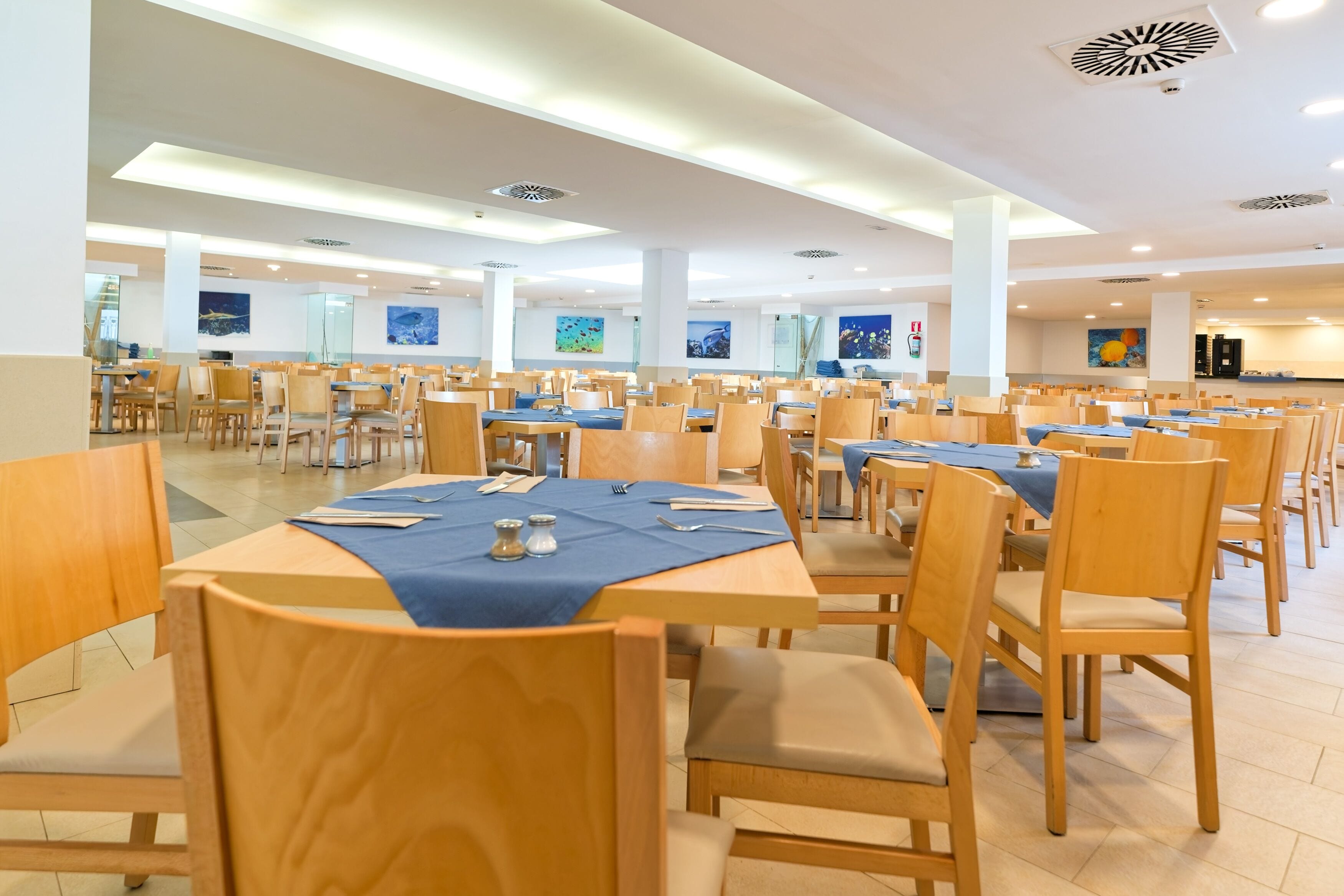 A spacious dining area with multiple tables and chairs, high ceilings with recessed lighting, and artwork on the walls.
