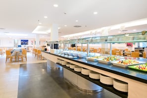 Zona de bufé con varias estaciones de comida, una larga barra con platos y un área de estar con sillas.