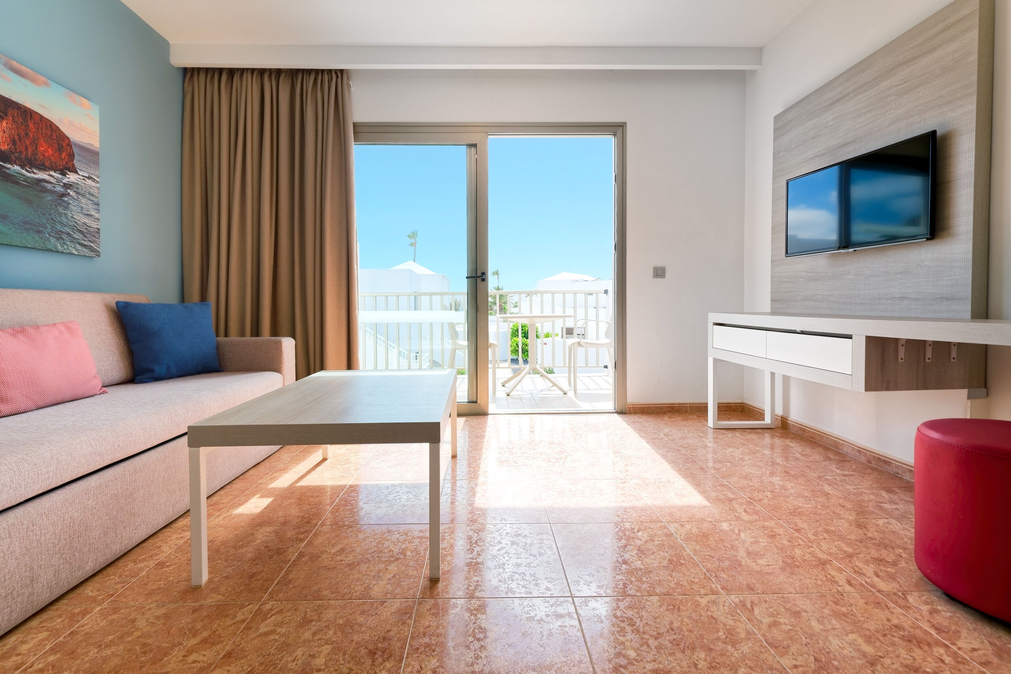 A modern hotel room with a sofa, a dining table, a TV mounted on the wall, and a balcony with umbrellas.