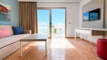 A modern hotel room with a sofa, a dining table, a TV mounted on the wall, and a balcony with umbrellas.