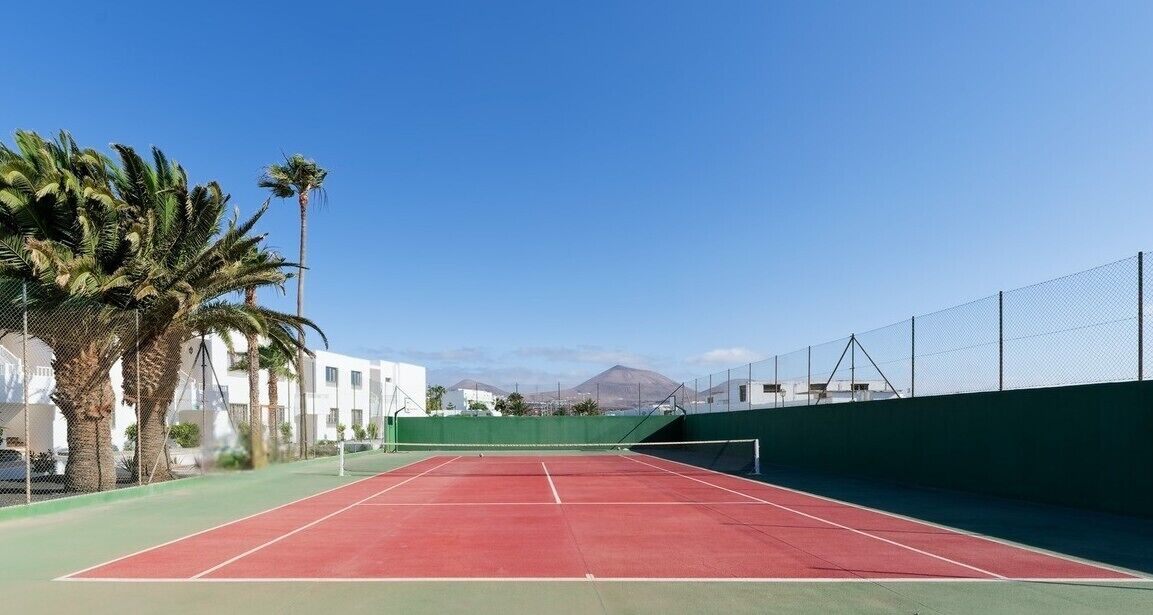 En tennisbane med net, omgivet af et grønt hegn, palmer og bygninger i baggrunden.
