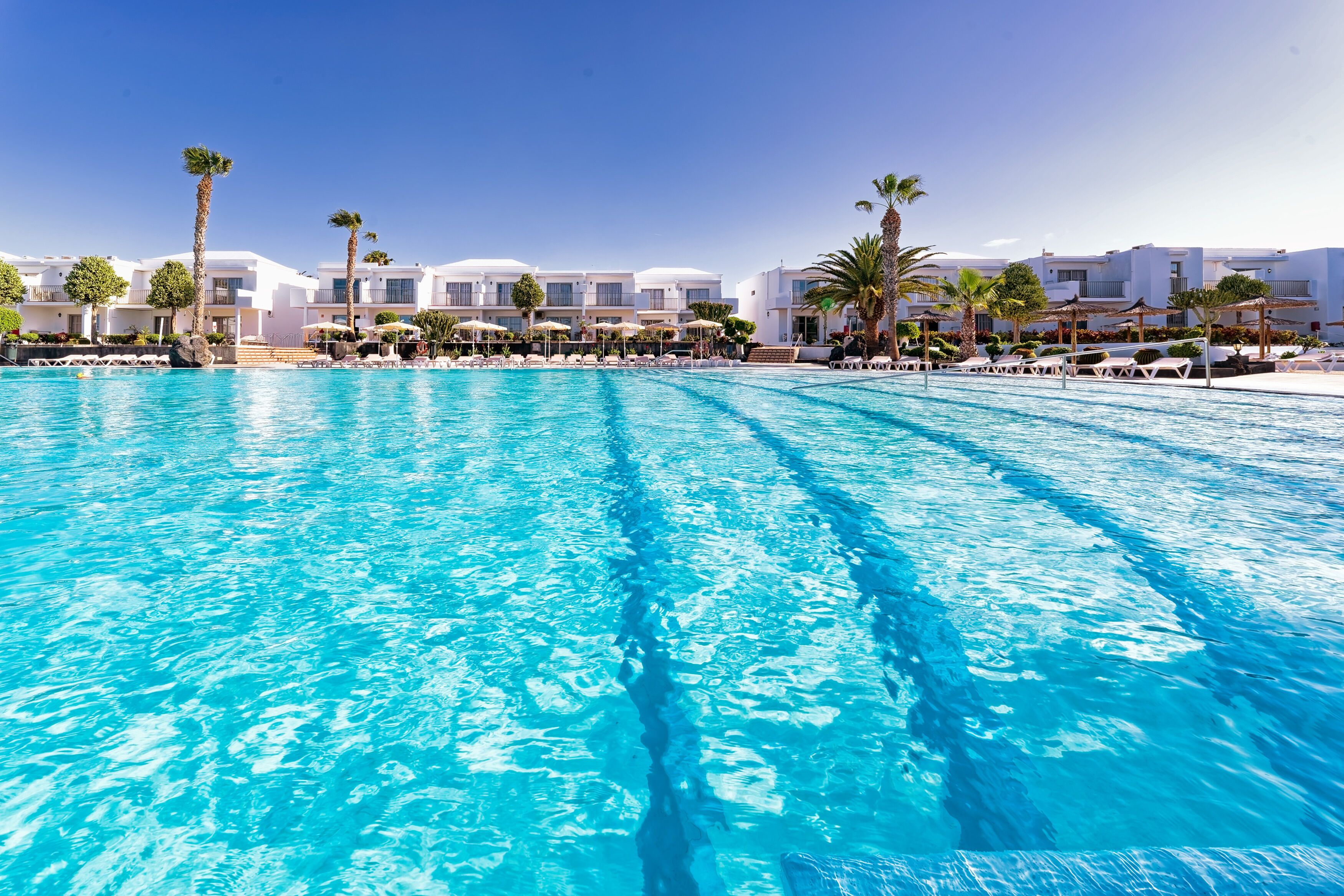 A large swimming pool with clear blue water, surrounded by white buildings with balconies and palm trees.