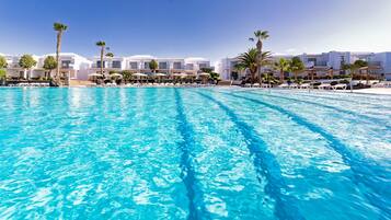 A large swimming pool with clear blue water, surrounded by white buildings with balconies and palm trees.