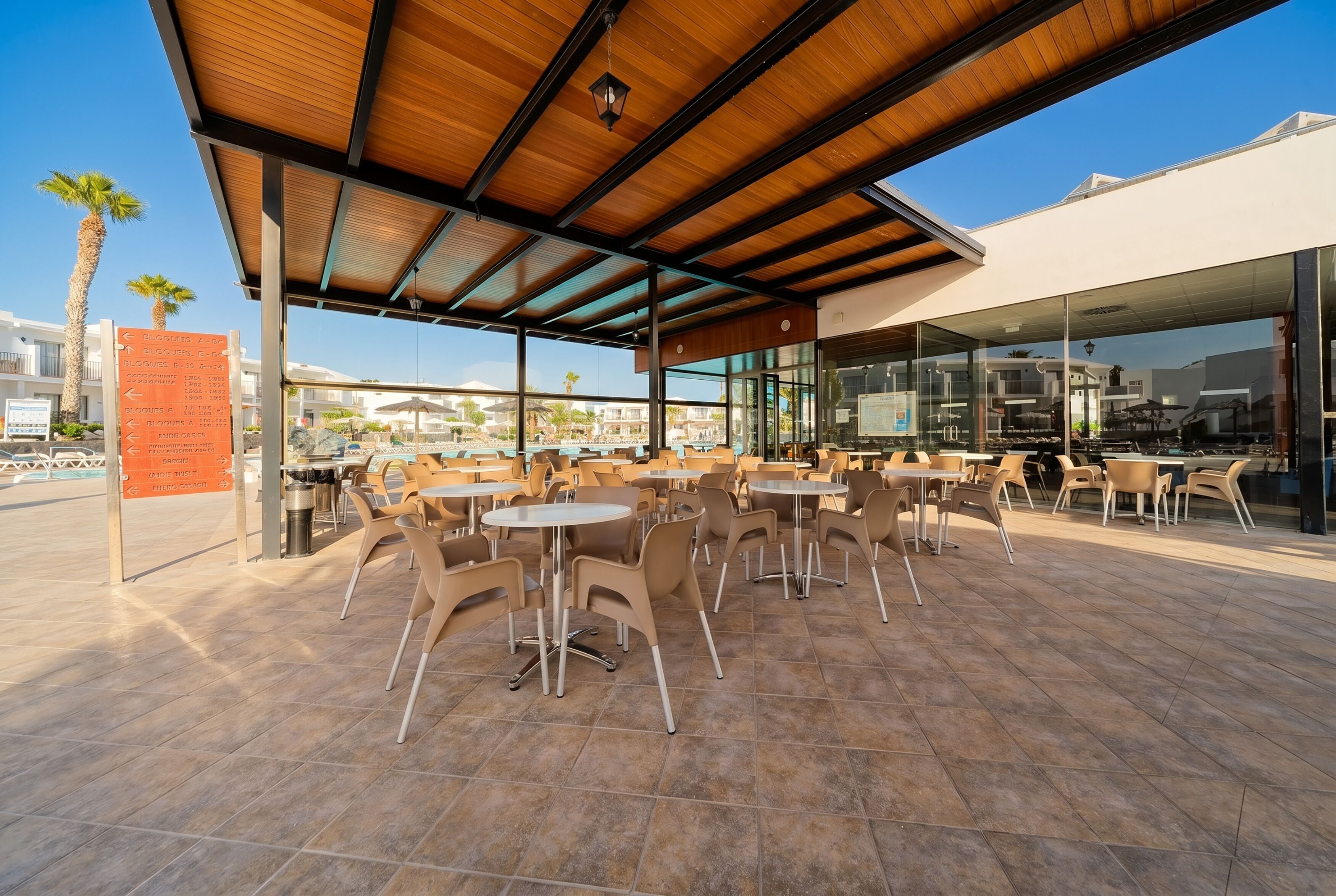 An outdoor seating area with tables and chairs, a covered section with a wooden roof, and a glass building in the background.