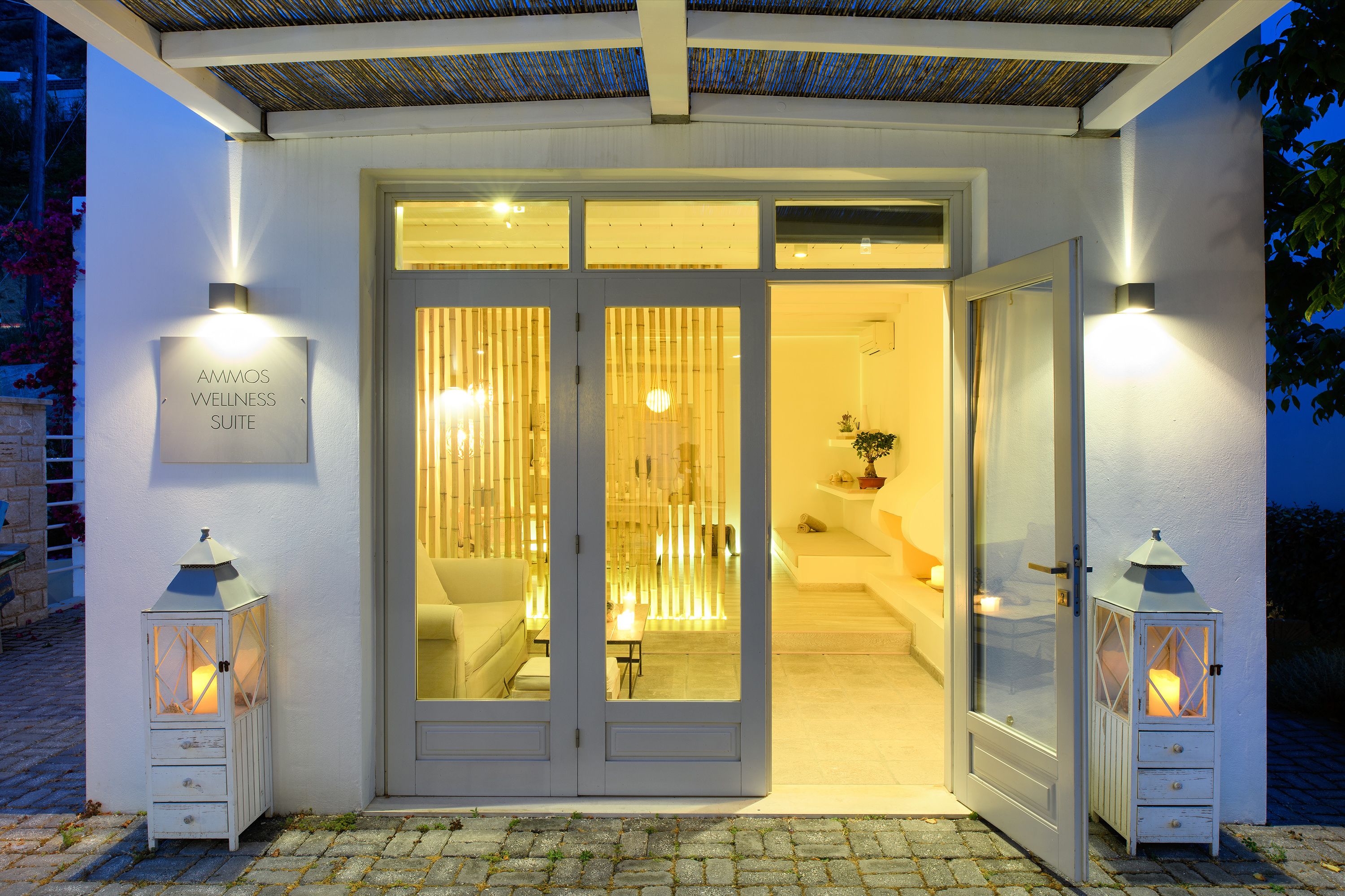 A well-lit entrance with a sign reading "AMMOS WELLNESS SUITE," a glass door, and two lantern-style lights.