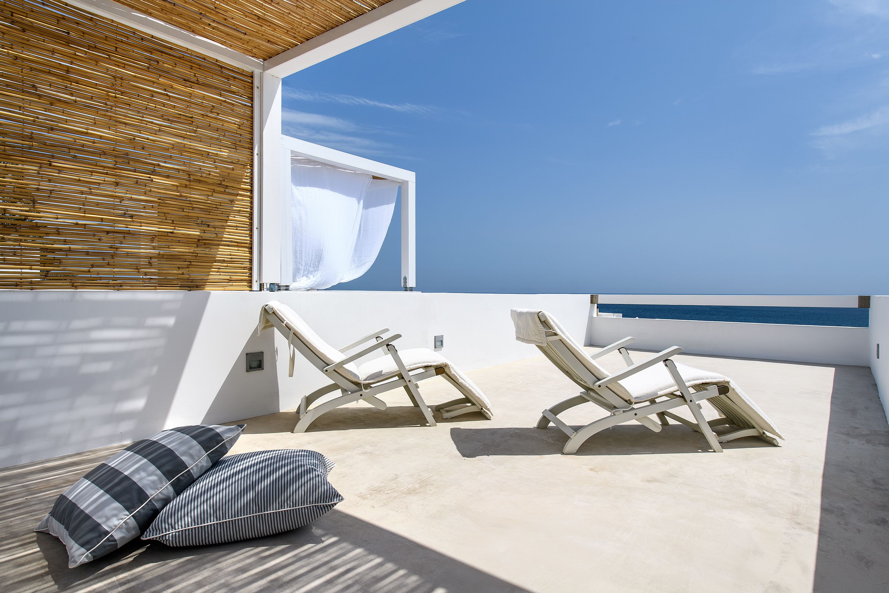 Terraza soleada con dos sillones de relax y un cojín, con vistas a un cielo azul y al mar.
