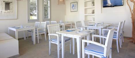 A dining area with white tables and chairs, a television mounted on the wall, and a large window with a view of the sea.