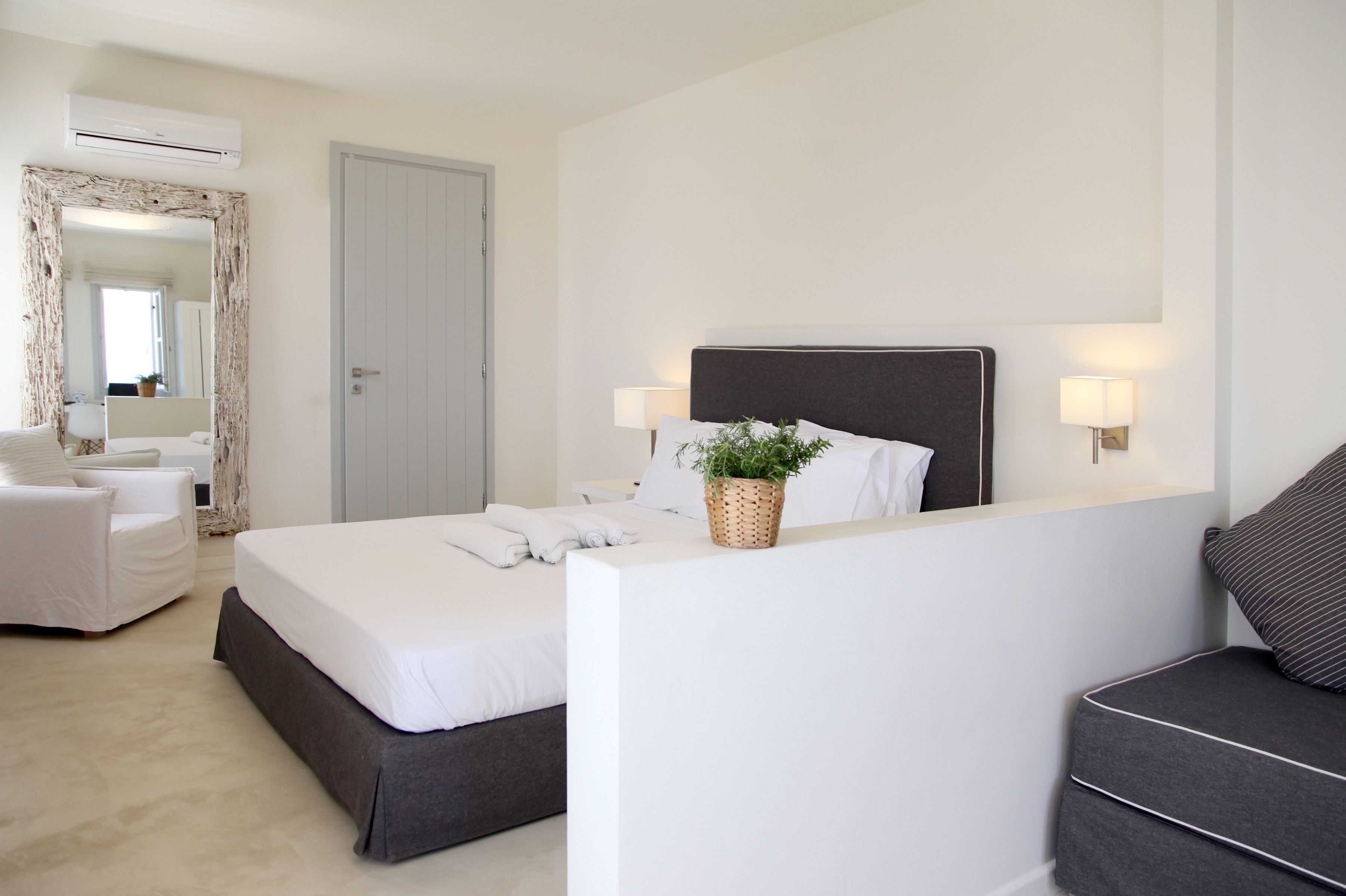 A modern hotel room with a large bed, a bedside table with a lamp, a chair, and a small table with a plant.