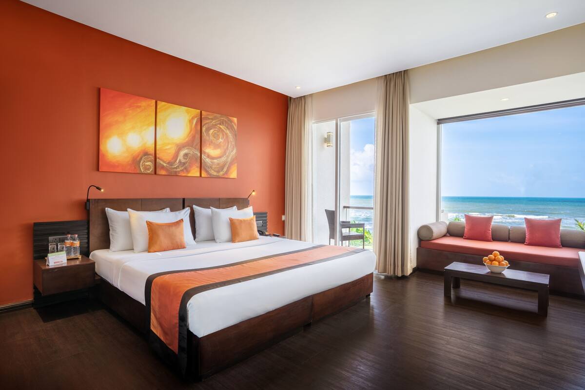 A hotel room with a large bed, a sofa, a view of the ocean, and artwork on the wall.