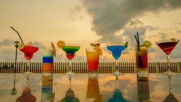 Vários cocktails coloridos sobre uma superfície refletora, com um pôr do sol ao fundo.