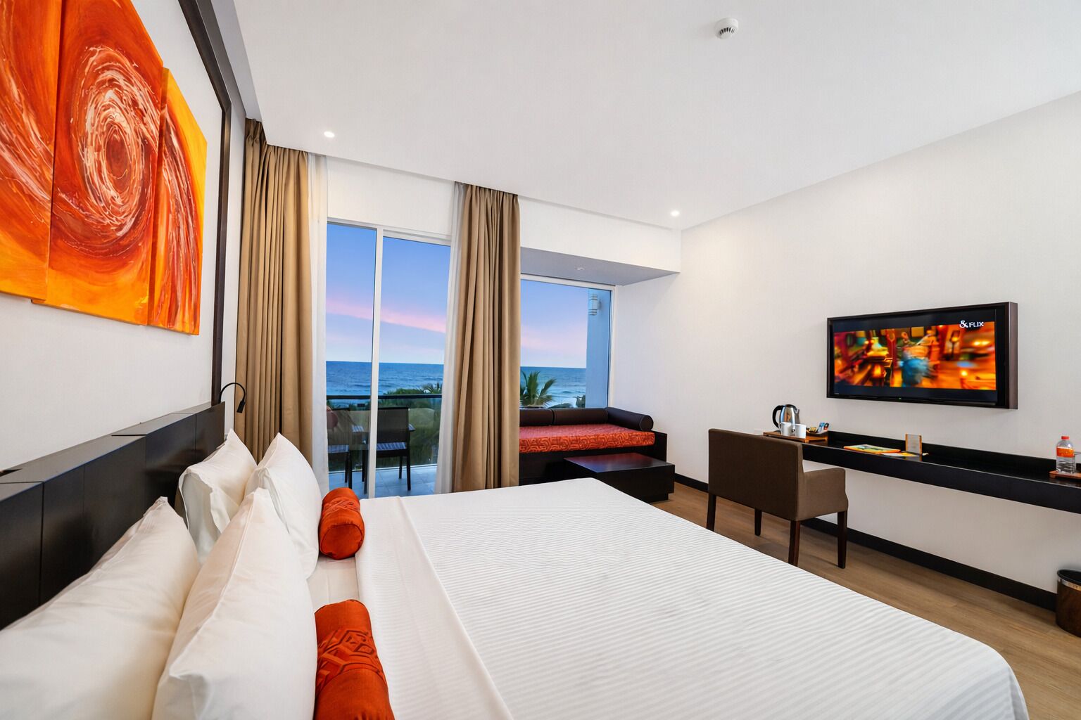 A hotel room with a large bed, a desk, a television, and a balcony with a view of the ocean.