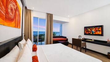 A hotel room with a large bed, a desk, a television, and a balcony with a view of the ocean.