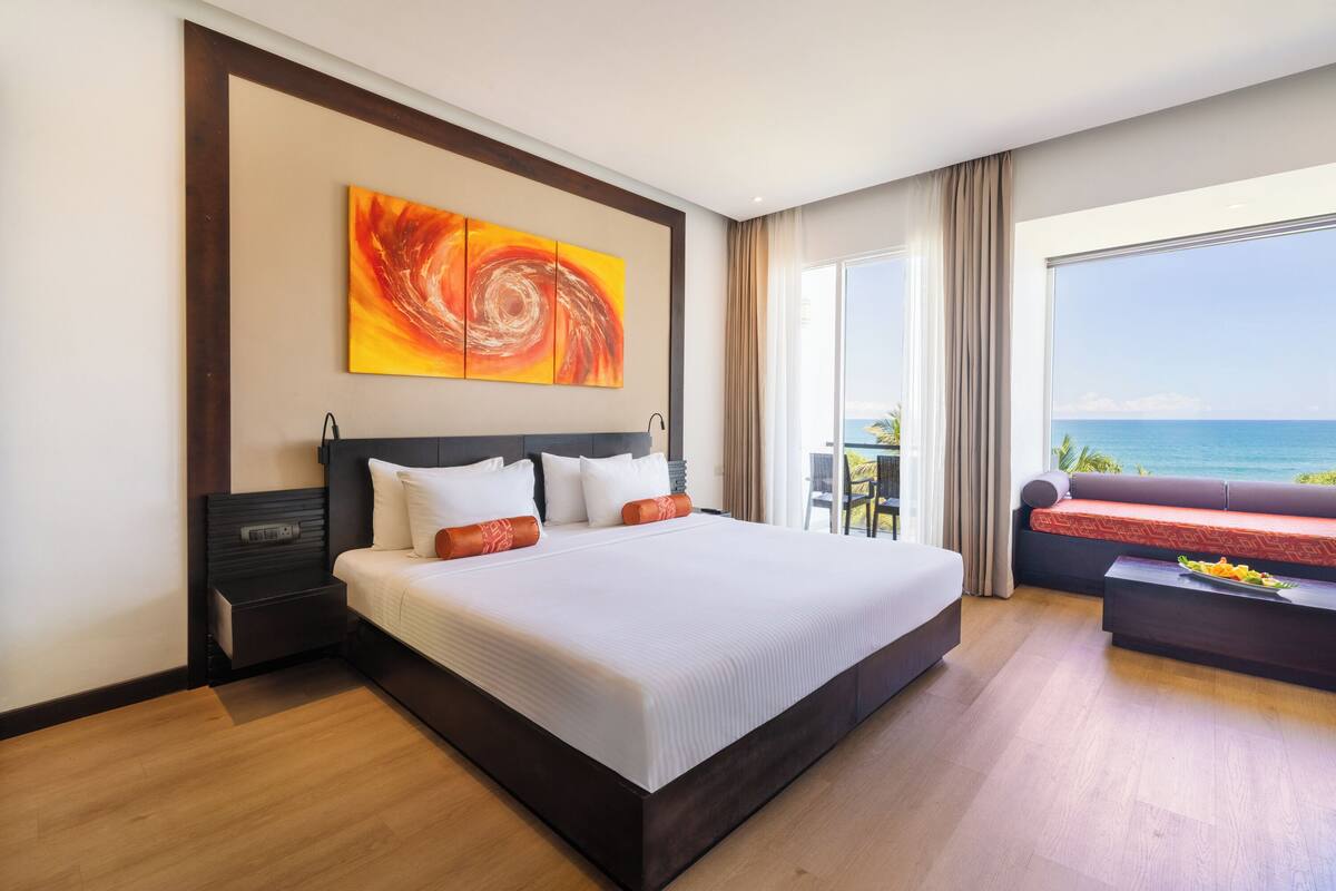 A hotel room with a large bed, a view of the ocean, a sofa, and a dining table.