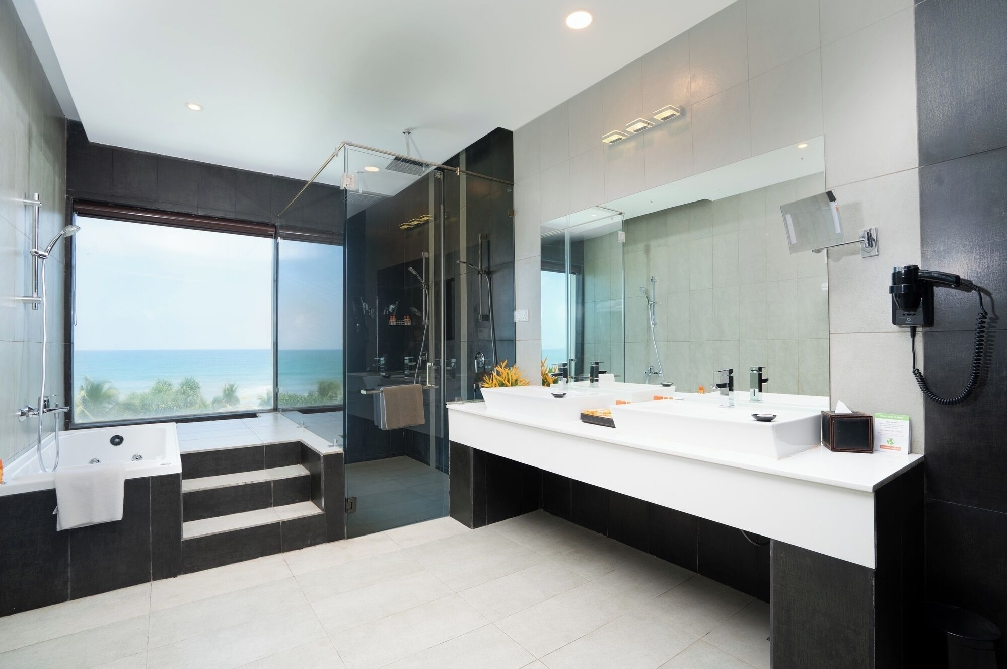 A modern bathroom with a large mirror, dual sinks, a glass shower enclosure, and a view of the ocean.