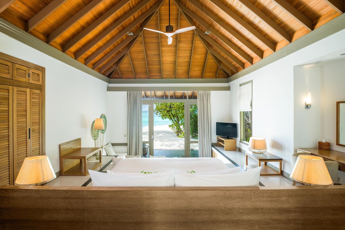 A hotel room with a wooden ceiling, a bed with white linens, a TV, a lamp, and a view of the beach through a glass door.
