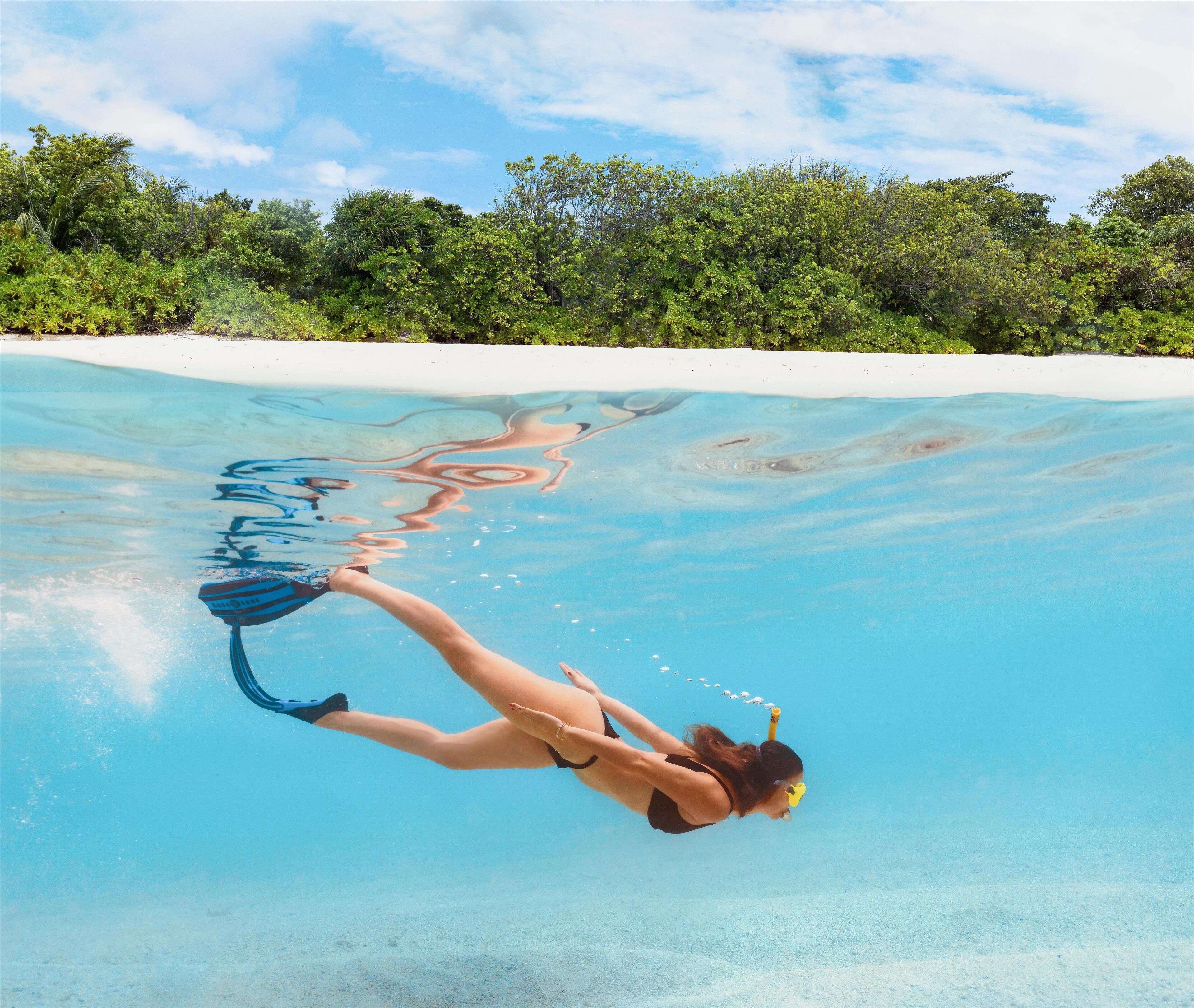 A person snorkeling in clear water with a sandy beach and lush greenery in the background.