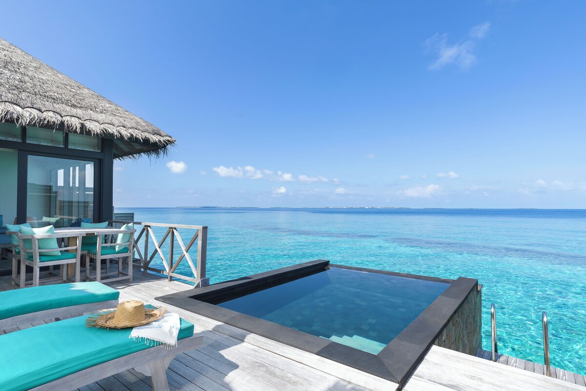 An infinity pool with a clear view of the ocean, wooden deck, lounge chairs, and a thatched-roof structure.