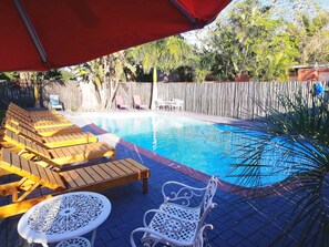 Une piscine entourée d’une clôture en bois et de plantes tropicales, avec des chaises longues en bois et des chaises en métal blanc.