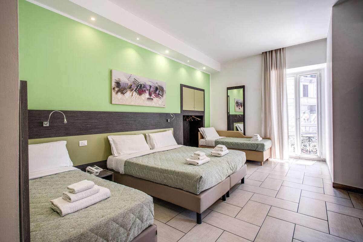 Habitación de hotel con tres camas, un ventanal con vistas y una pared verde.