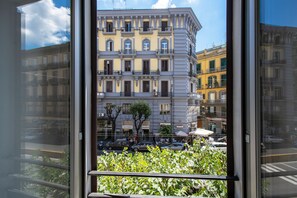 A view through a window of a multi-story building with balconies, a street with cars, and a clear sky. - Napoli Suite (Naples)
