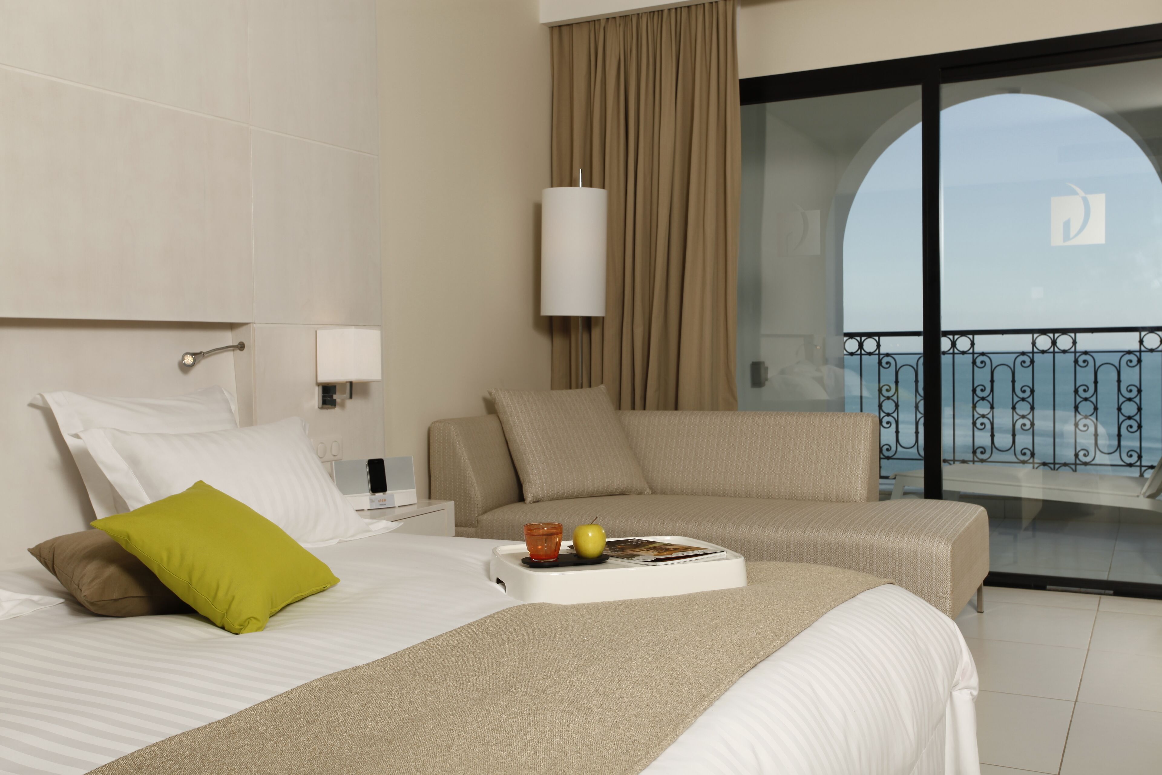 A hotel room with a bed, two sofas, a balcony with a view, and a table with a tray.