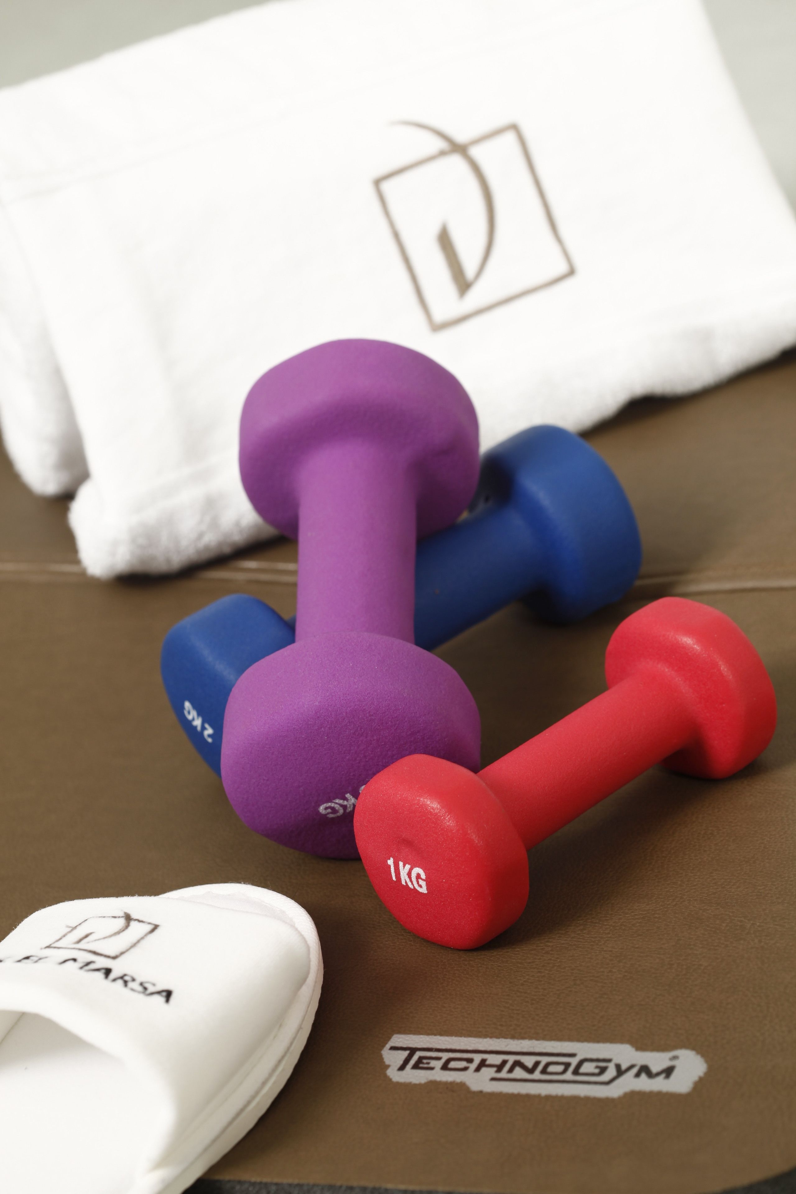 A pair of dumbbells, a white towel, and white slippers on a brown surface with a logo.