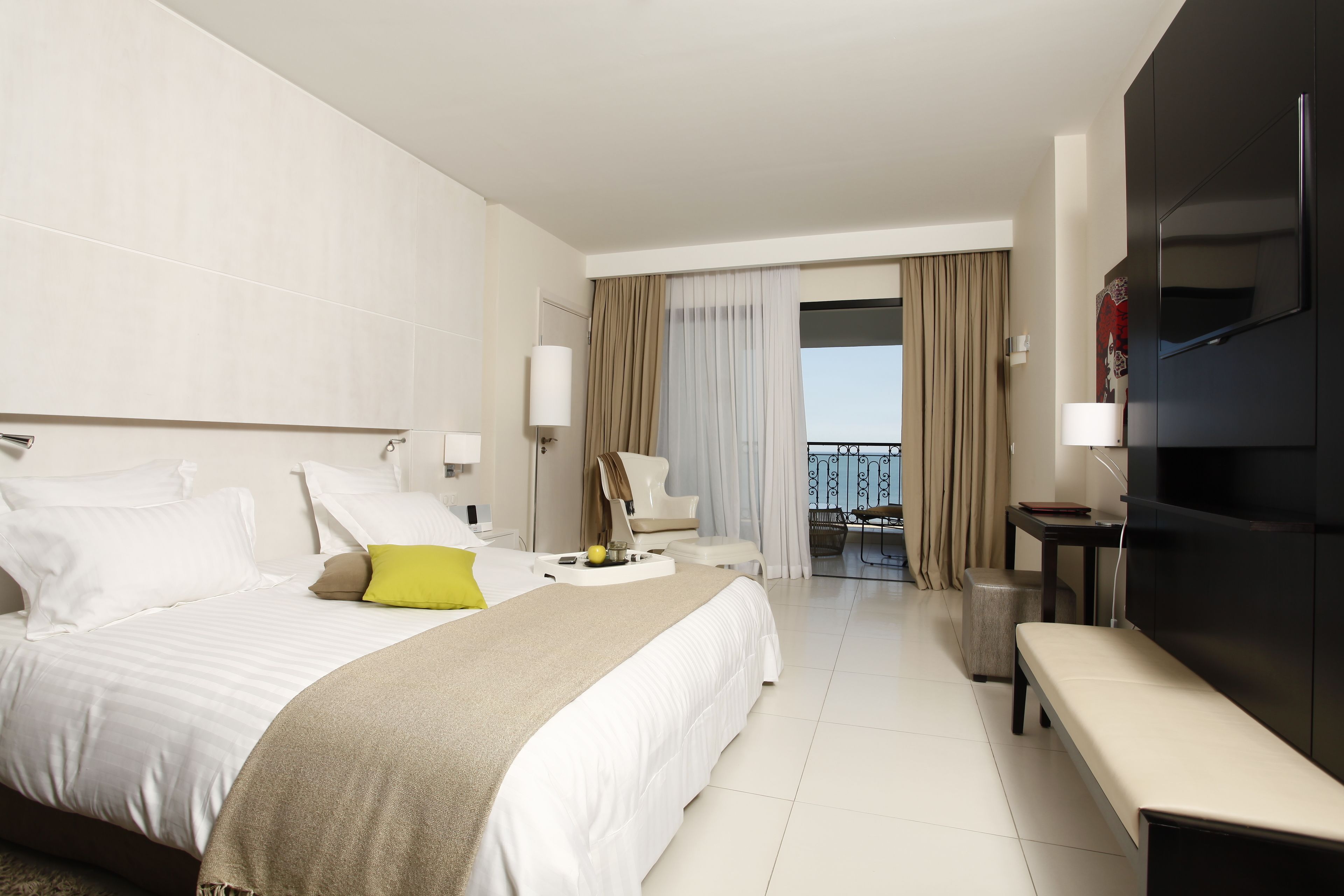 Una habitación de hotel moderna con una cama grande, un banco, un televisor y un balcón con cortinas.