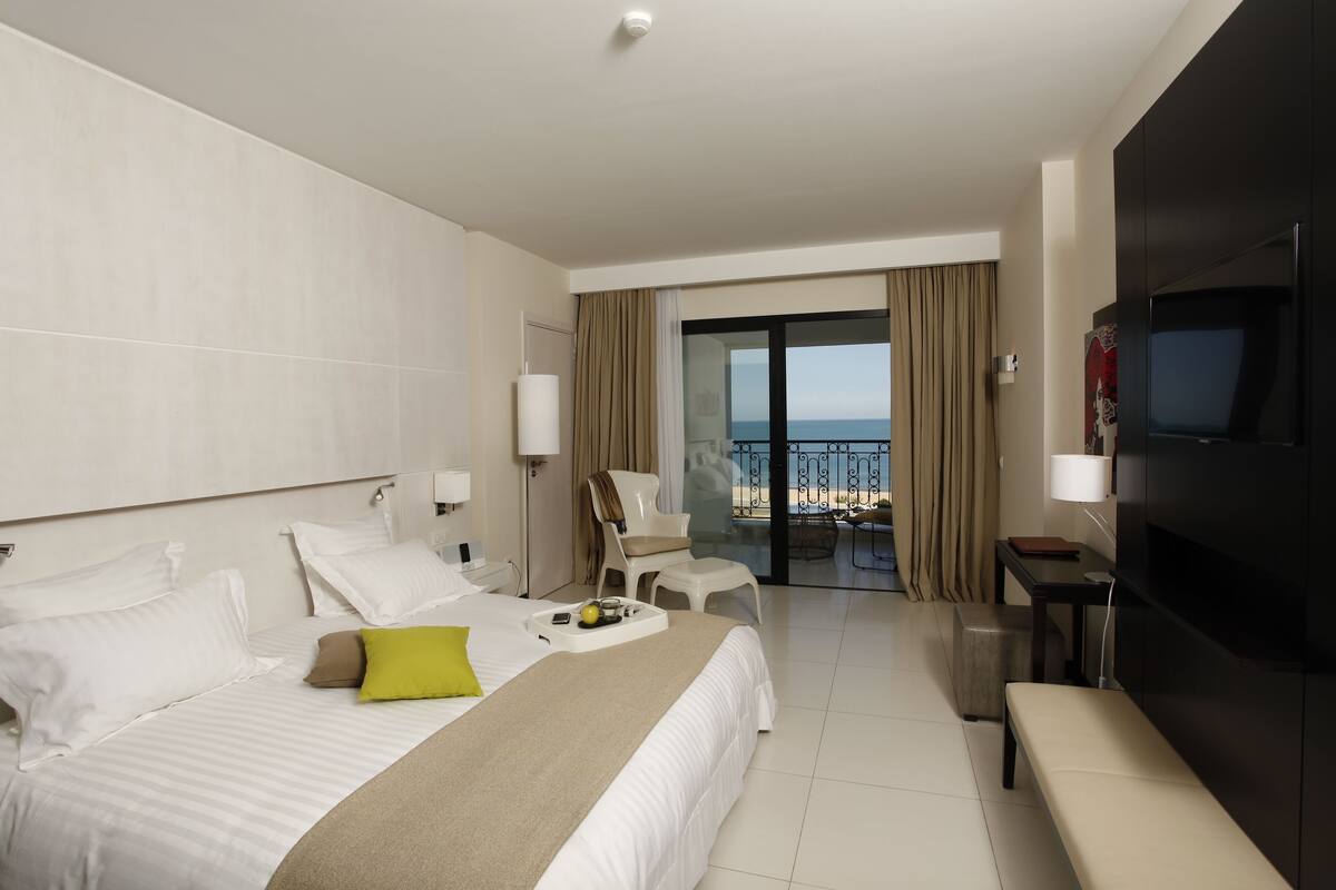 Habitación de hotel con cama, banca, televisor, escritorio y vista a la playa.