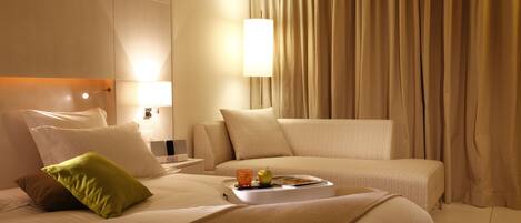 A hotel room with a bed, a nightstand, a lamp, a tray with a glass and an apple, and beige curtains.