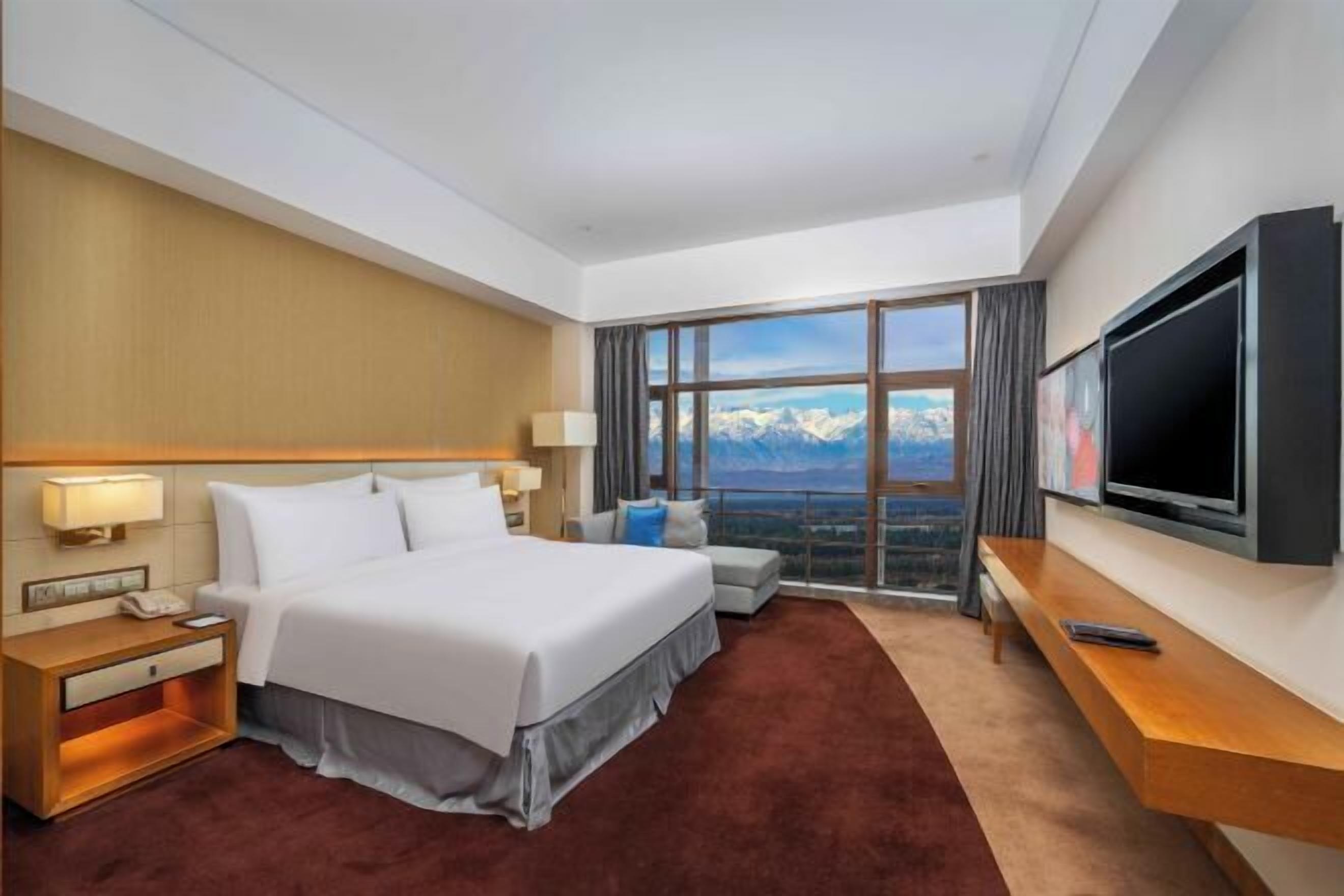 A hotel room with a large bed, a TV, a desk, and a view of mountains.