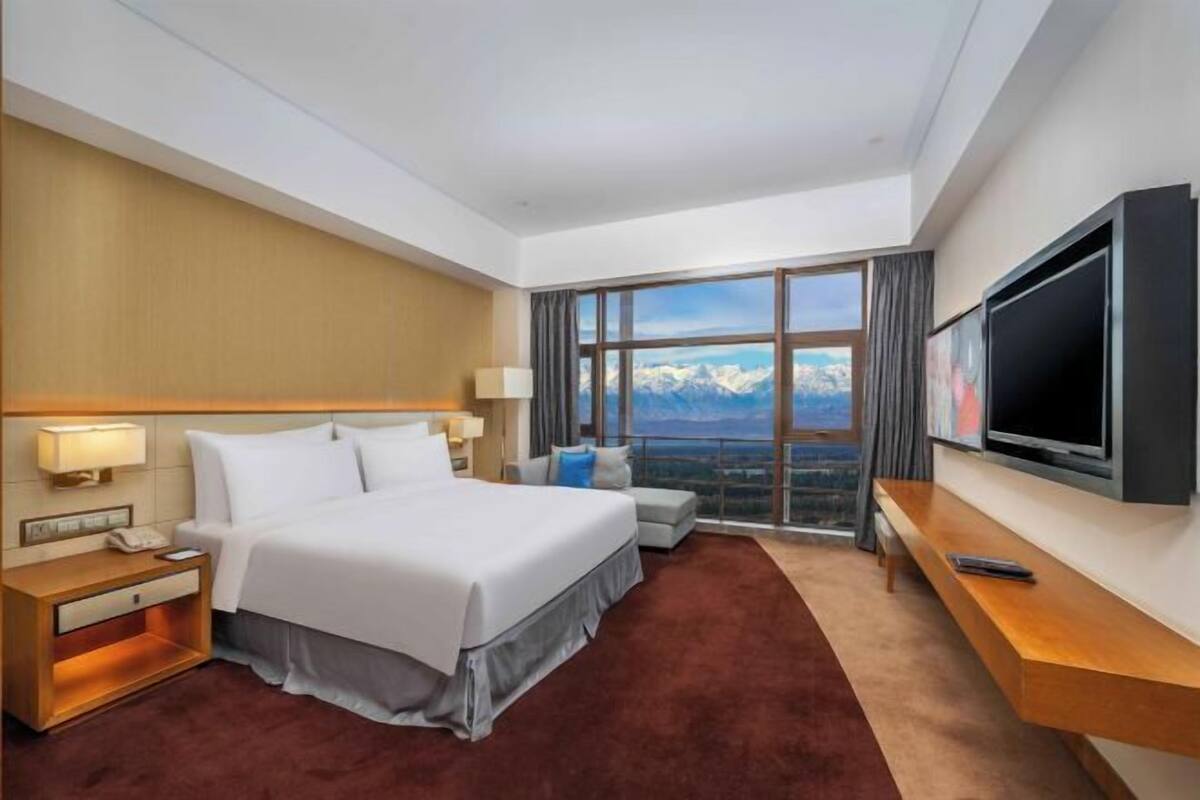 A hotel room with a large bed, a TV, a desk, and a view of mountains.