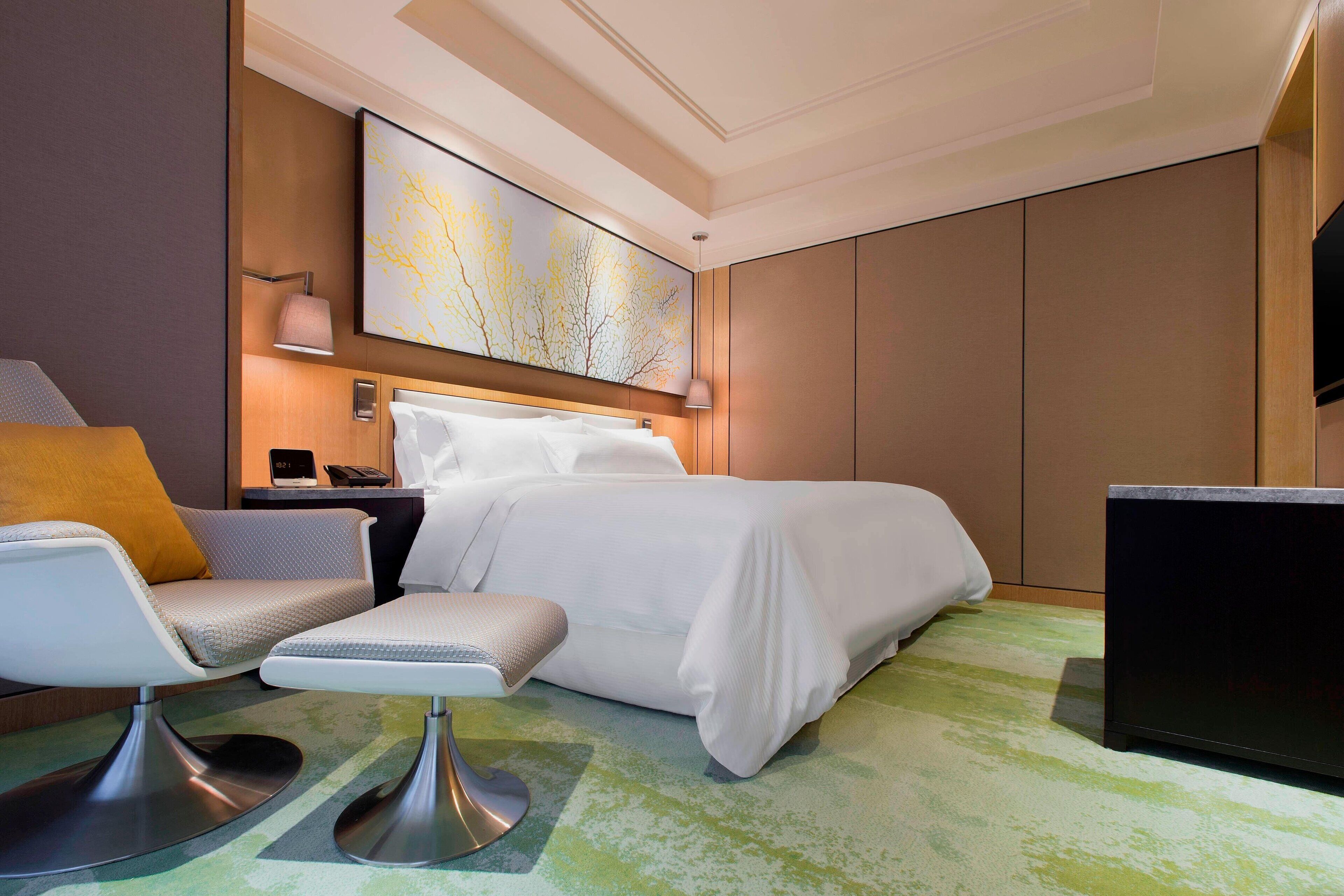 A modern hotel room with a bed, a seating area with a chair and ottoman, a desk, and a large abstract painting on the wall.
