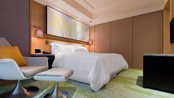 A modern hotel room with a bed, a seating area with a chair and ottoman, a desk, and a large abstract painting on the wall.