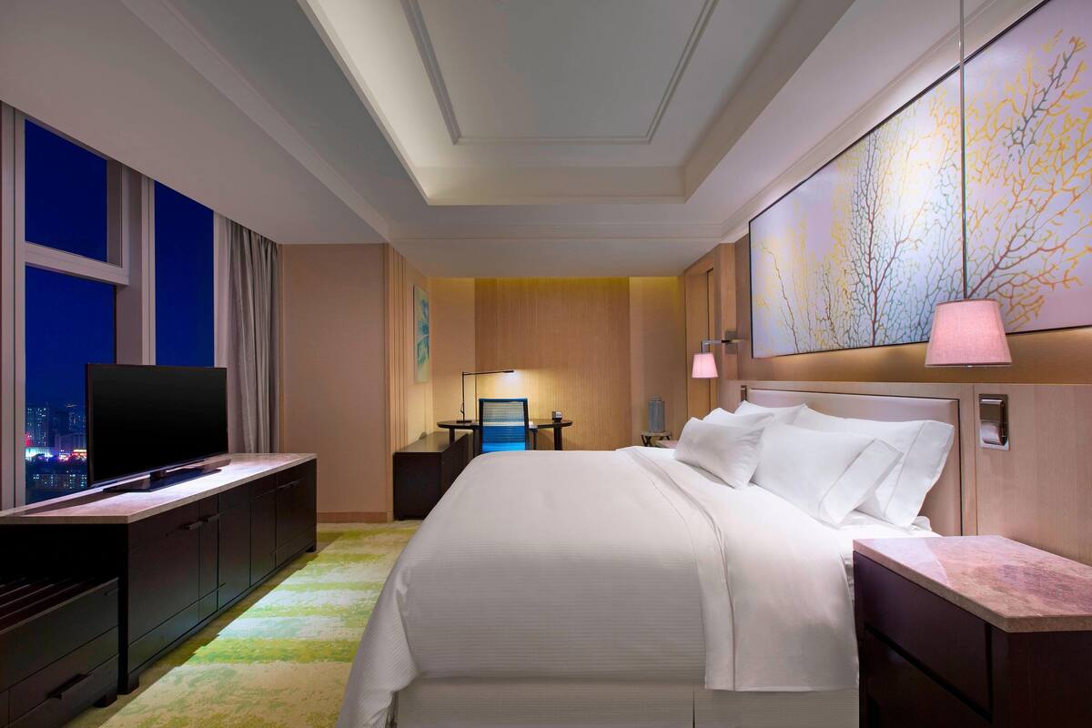 Une chambre d’hôtel avec un grand lit, un téléviseur posé sur une commode, un bureau avec une chaise et une vue sur la ville par la fenêtre.