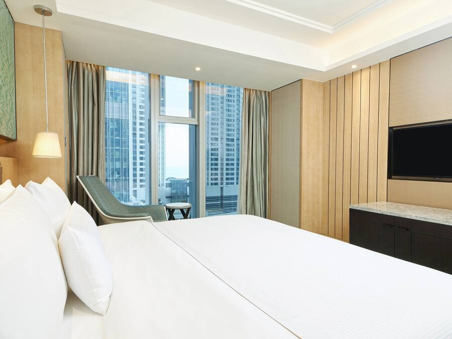 Quarto de hotel com uma cama grande, televisão na parede, escrivaninha com cadeira e vista para edifícios altos pela janela.