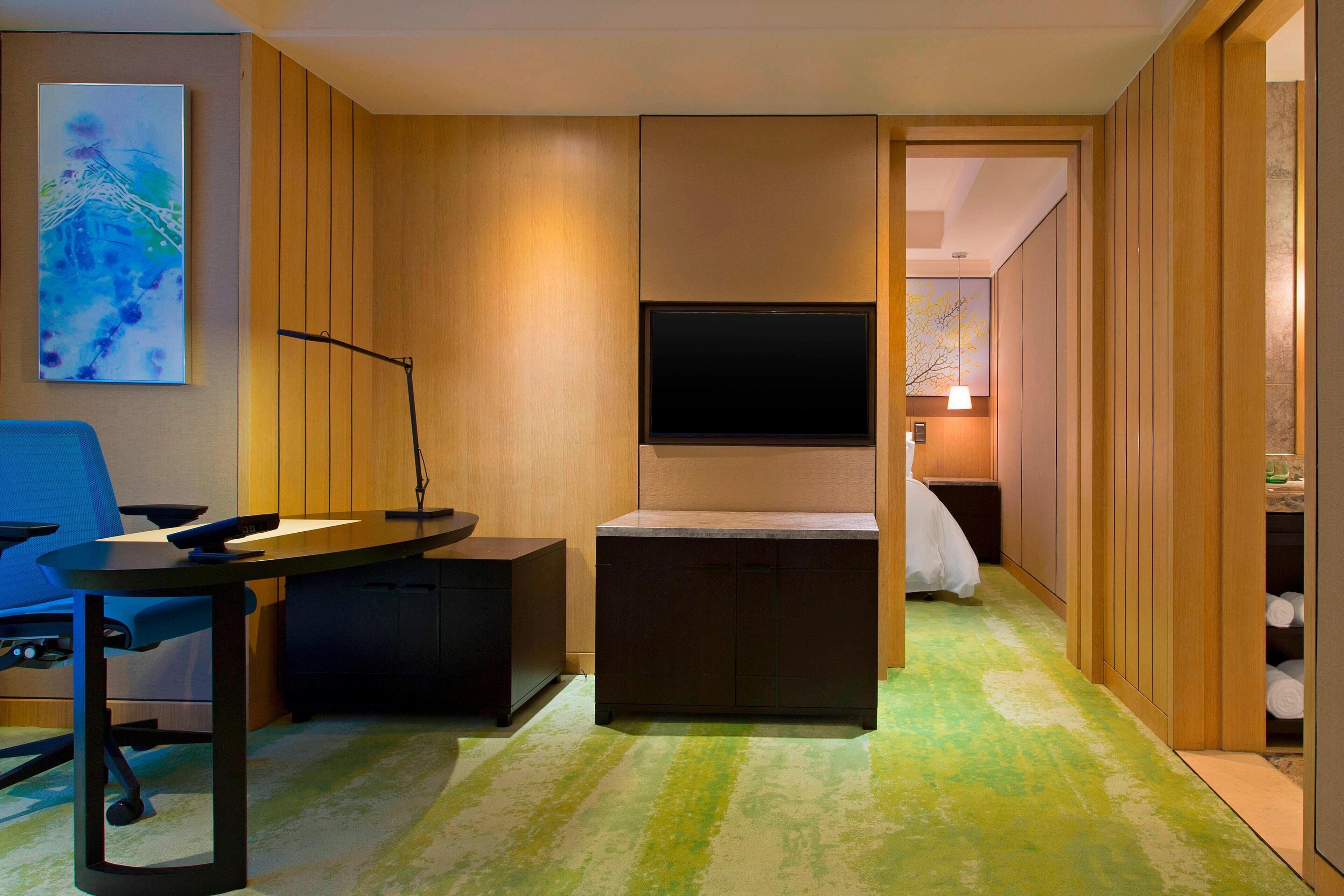 A modern hotel room with a flat-screen TV mounted on the wall, a desk with a chair, and a bed with a nightstand.