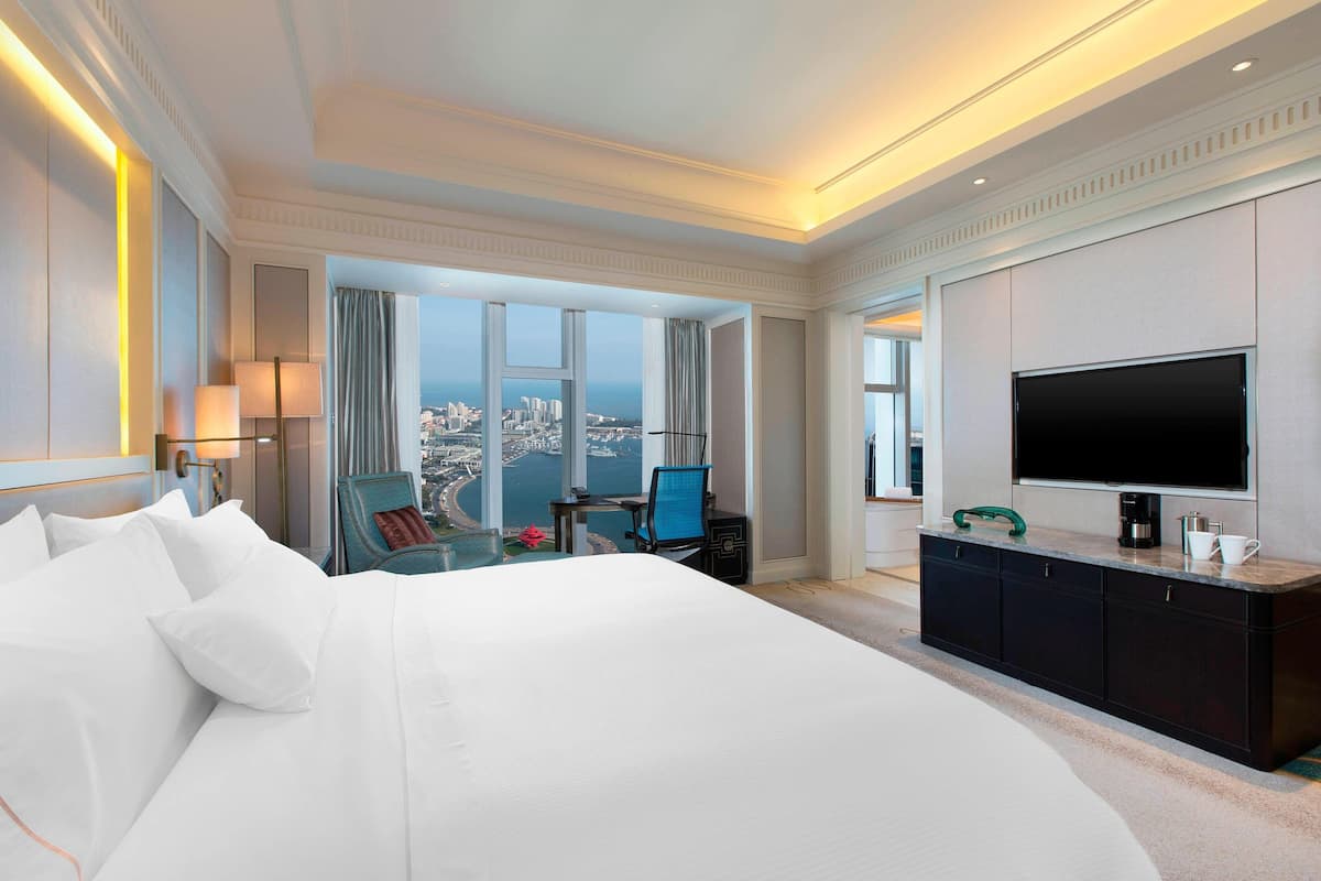 Une chambre d’hôtel avec un grand lit, un bureau avec une chaise, une télévision et une vue sur le paysage urbain grâce à de grandes fenêtres.