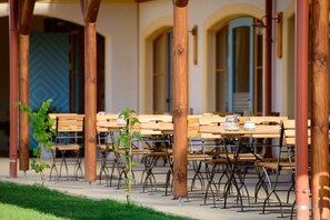 An outdoor seating area with wooden tables and chairs, a grassy foreground, and a building with arched windows in the background.
