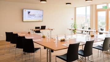 A conference room with a large table, black chairs, a screen displaying a presentation, and a view of the outdoors through large windows.