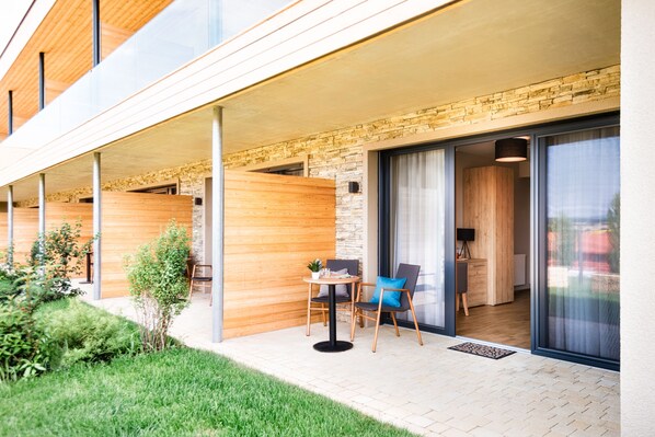 A modern building with a covered outdoor area featuring a wooden deck, a small table with chairs, and a view into the interior. - JUFA Hotel Neutal (Neutal)
