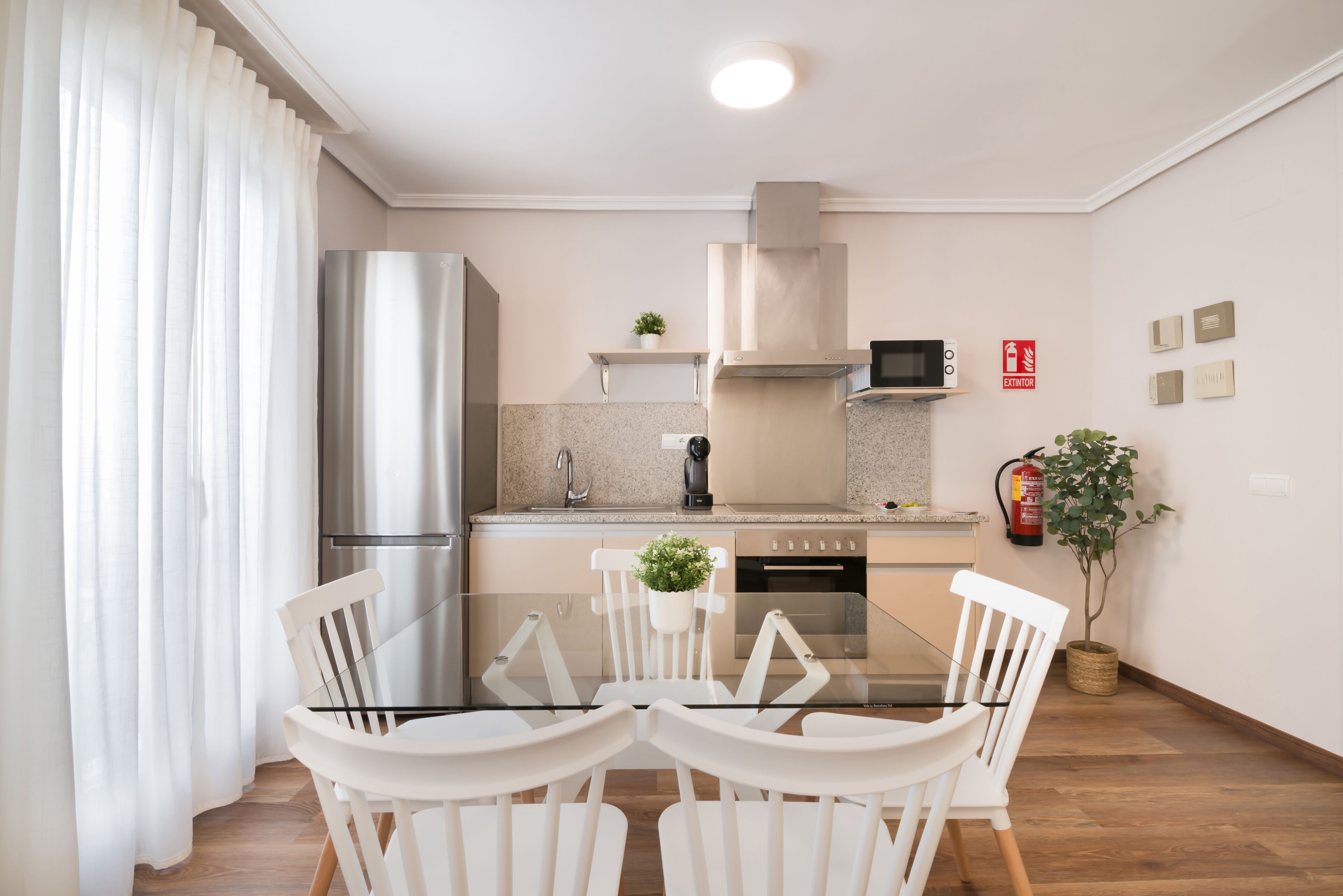 A modern kitchen with a glass dining table, white chairs, stainless steel appliances, and a fire extinguisher on the wall.
