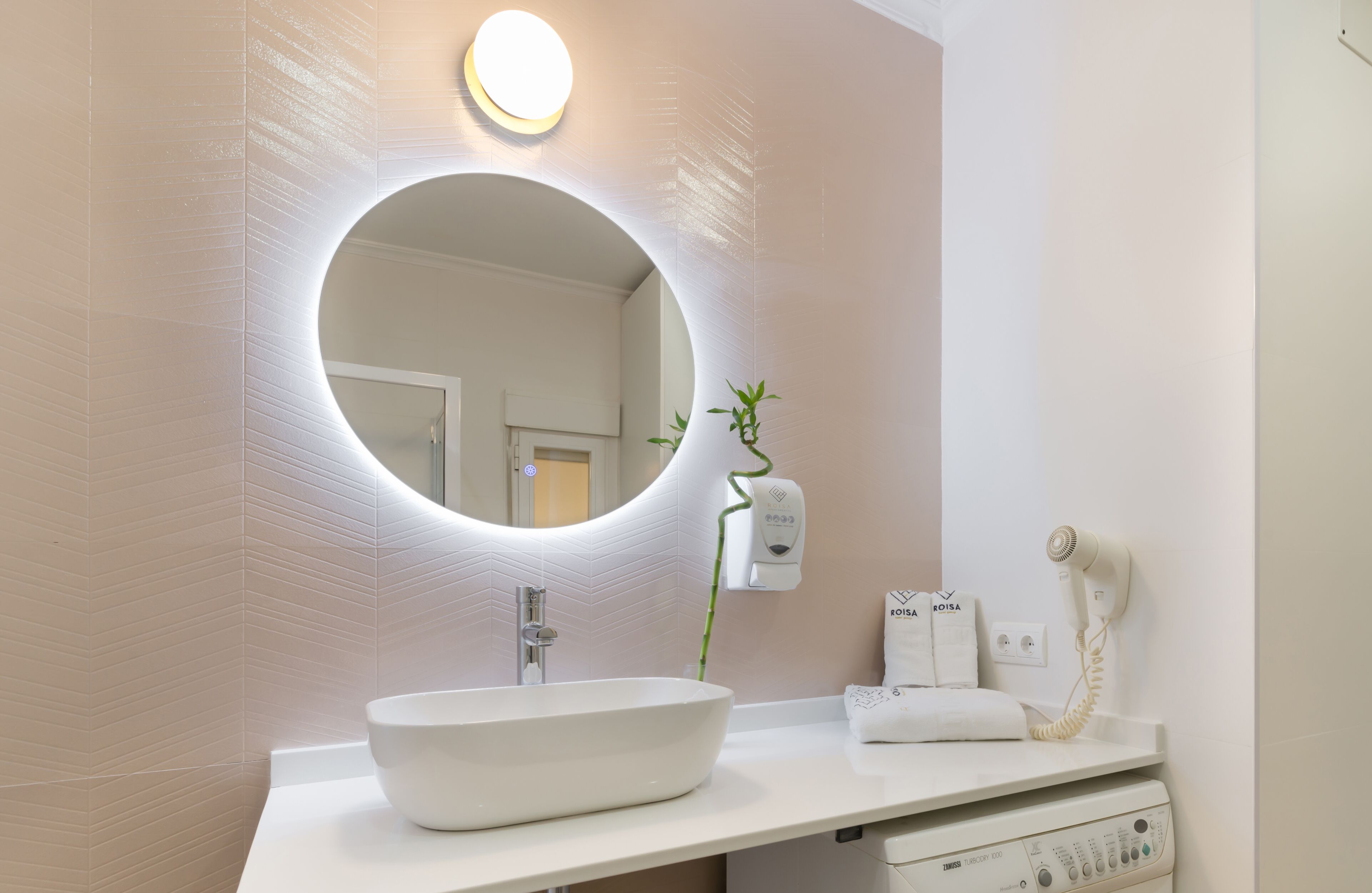 Un bagno moderno con uno specchio tondo, un lavandino bianco, un asciugacapelli portatile e un dispenser di sapone per le mani fissato alla parete.