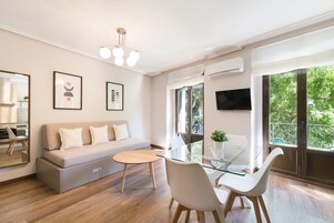 A modern living room with a sofa, glass dining table, and chairs, a TV, and large windows with a view of greenery.