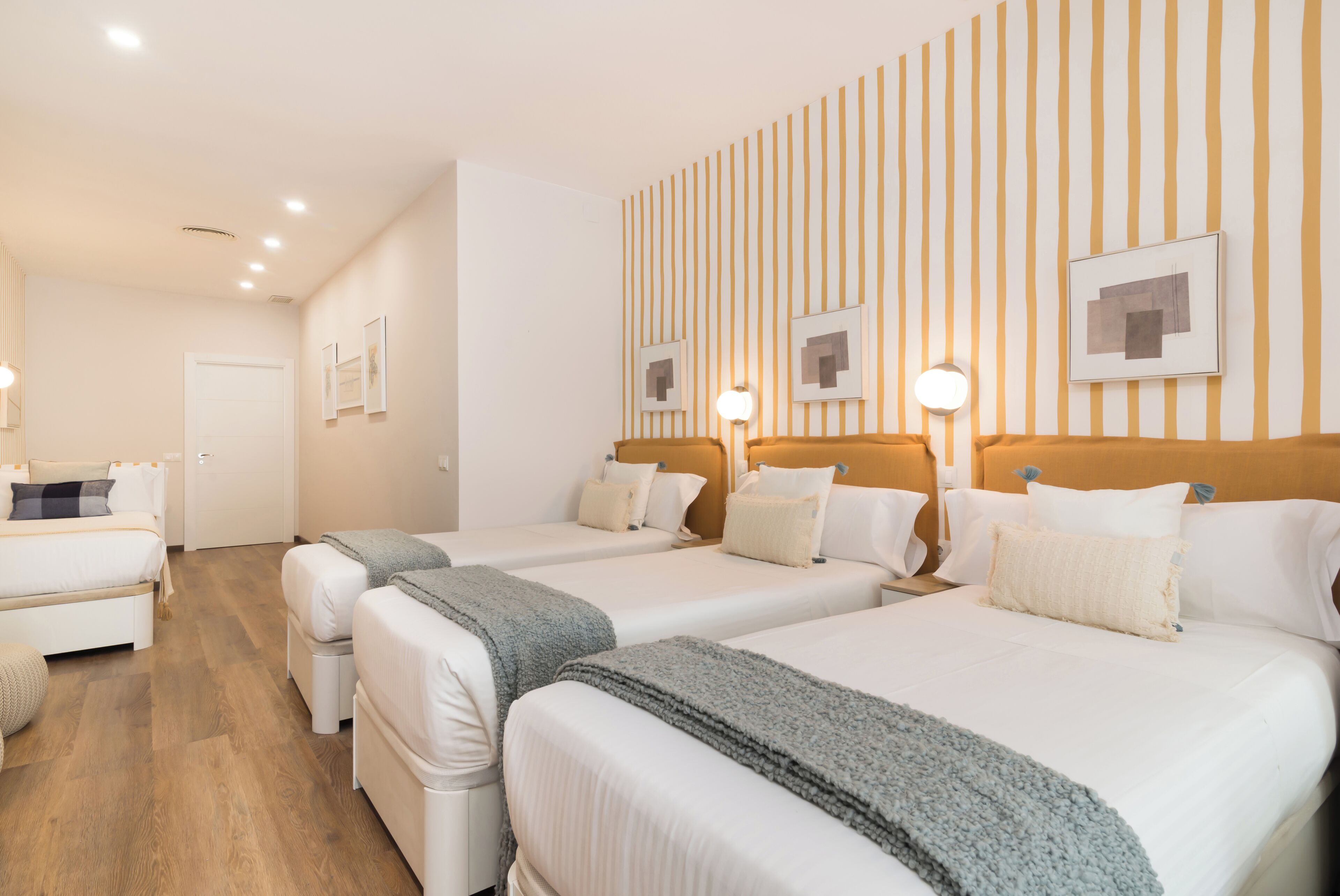 Habitación de hotel con cuatro camas dispuestas en fila, suelo de madera y pared a rayas con cuadros enmarcados.