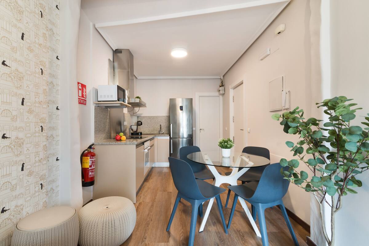 A compact kitchen with modern appliances, a dining area with blue chairs, and a fire extinguisher on the wall.